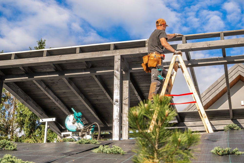 A worker stands on a ladder, expertly installing solar panels on a rooftop, surrounded by trees and a clear blue sky, during daylight hours.
