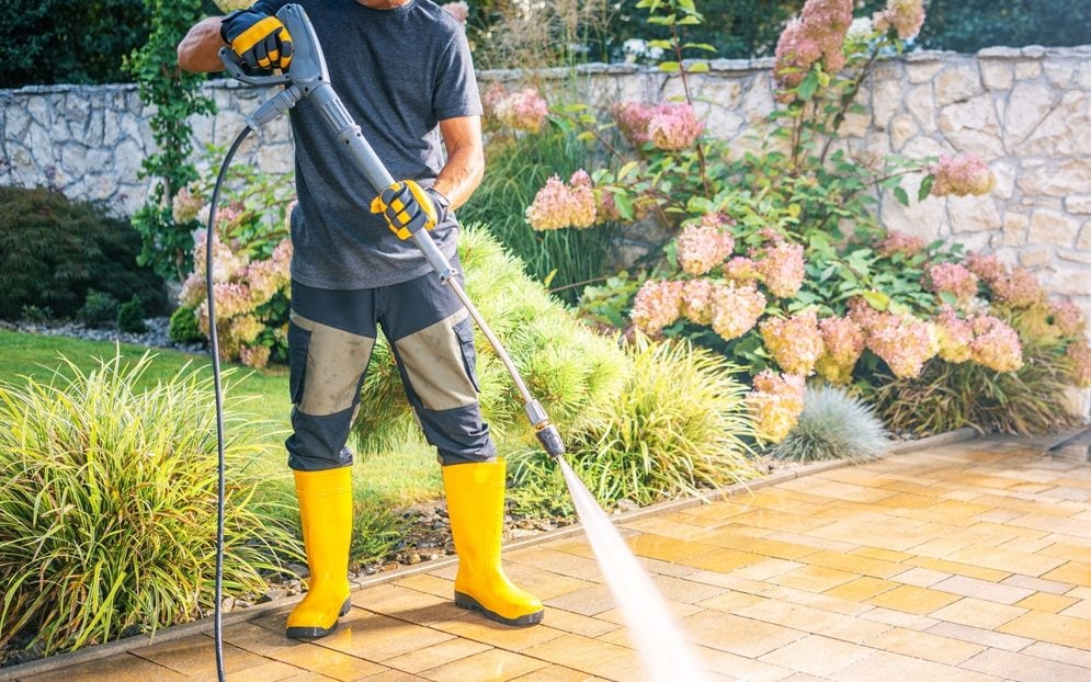 A person wearing yellow boots and gloves operates a pressure washer while cleaning a paved patio in a lush garden