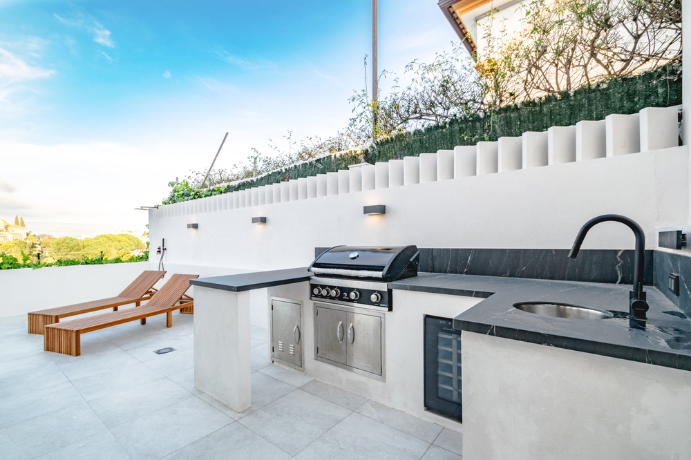 Outdoor Kitchen on a Sunlit Patio with Built‑in Gas Grill, Sink, Counter, and Mini Fridge Beside Two Wooden Lounge Chairs Under a White Wall.
