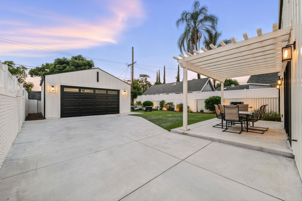 a Modern Backyard with a Concrete Patio, White Pergola, and a Sleek Black and White Detached Garage at Sunset.