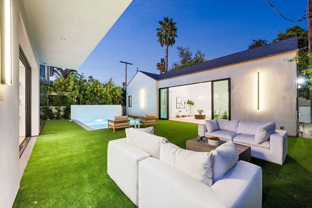 Modern backyard with white outdoor sofas on green artificial grass, a rectangular pool, and a contemporary white house with sliding glass doors at dusk/early evening