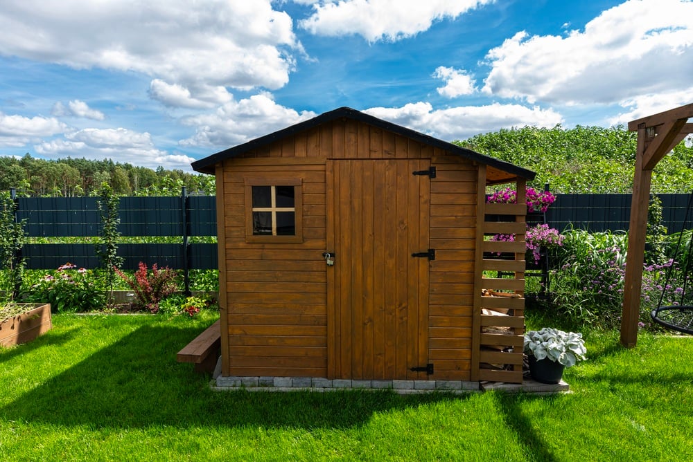 Brown wooden garden shed with a small window and padlock, set on a green lawn in a sunny backyard.
