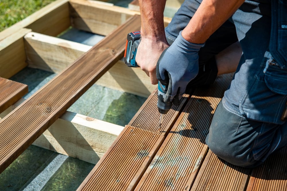 Person wearing gloves drills a wooden deck plank with a cordless drill at a construction site